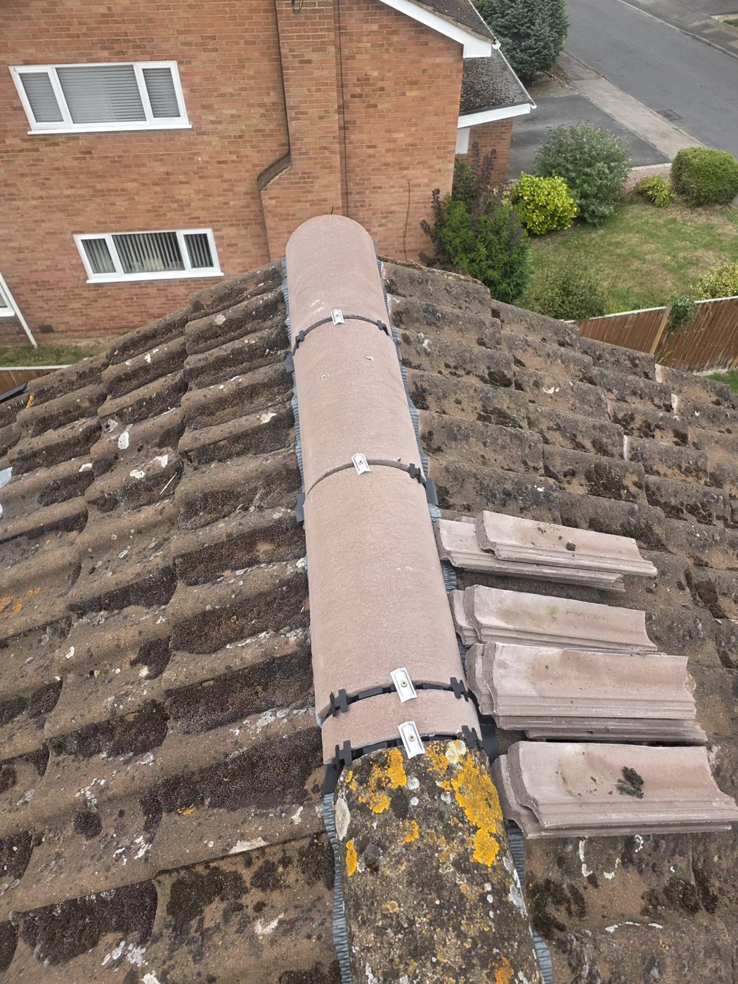New ridge tiles clipped along a roof next to an old lichen-covered ridge tile, with spare tiles stacked on the roof