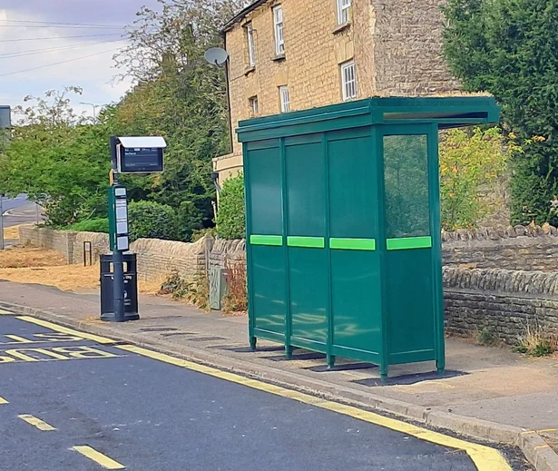 A new dark green bus shelter with a lime green band, beside a stone wall on Oxford Hill, Witney, with a bus stop pole and bin next to it