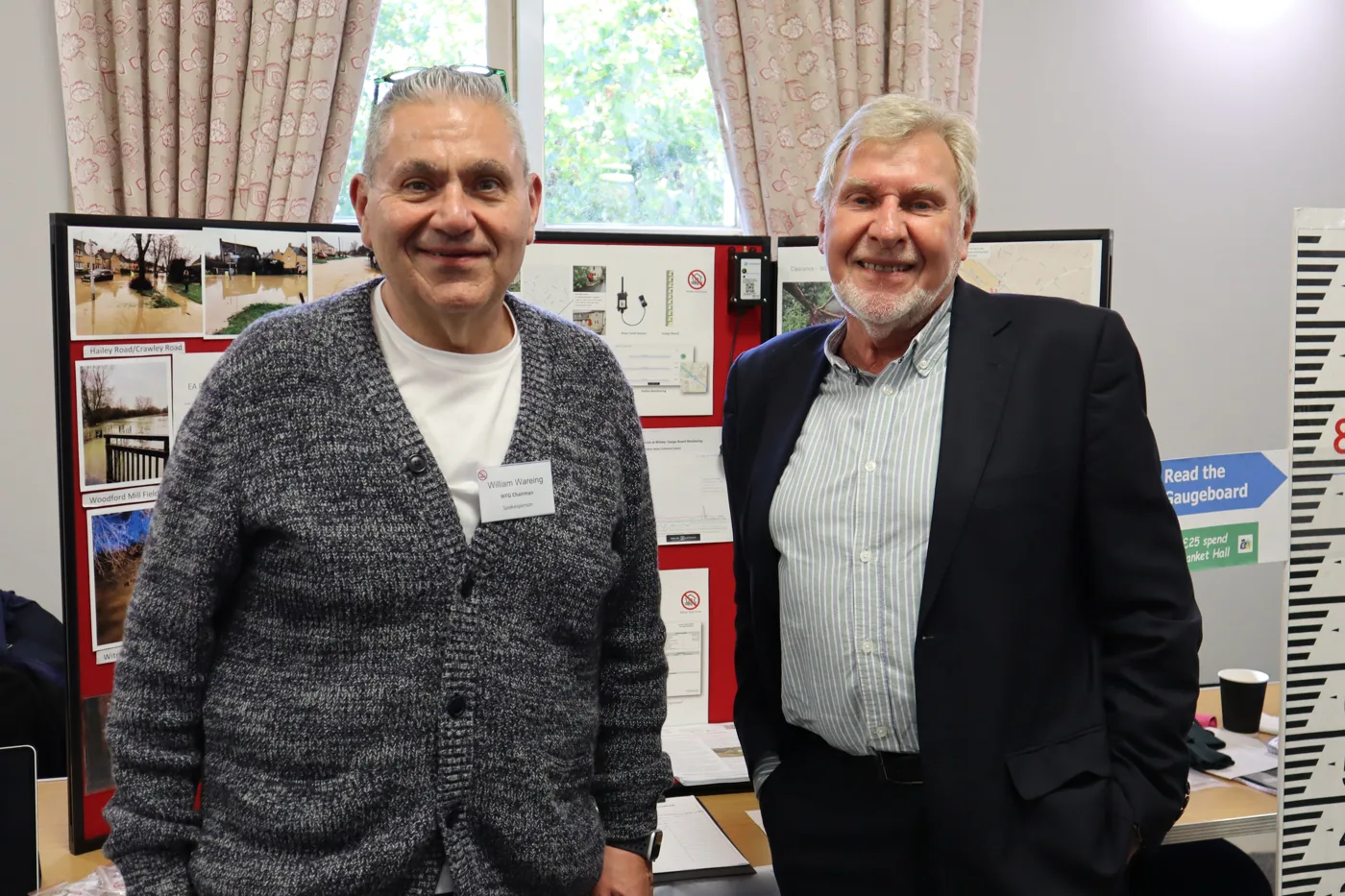Two men smiling in front of a Witney Flood Group display board of flood photographs, a river gauge sensor and a tall depth gauge marker