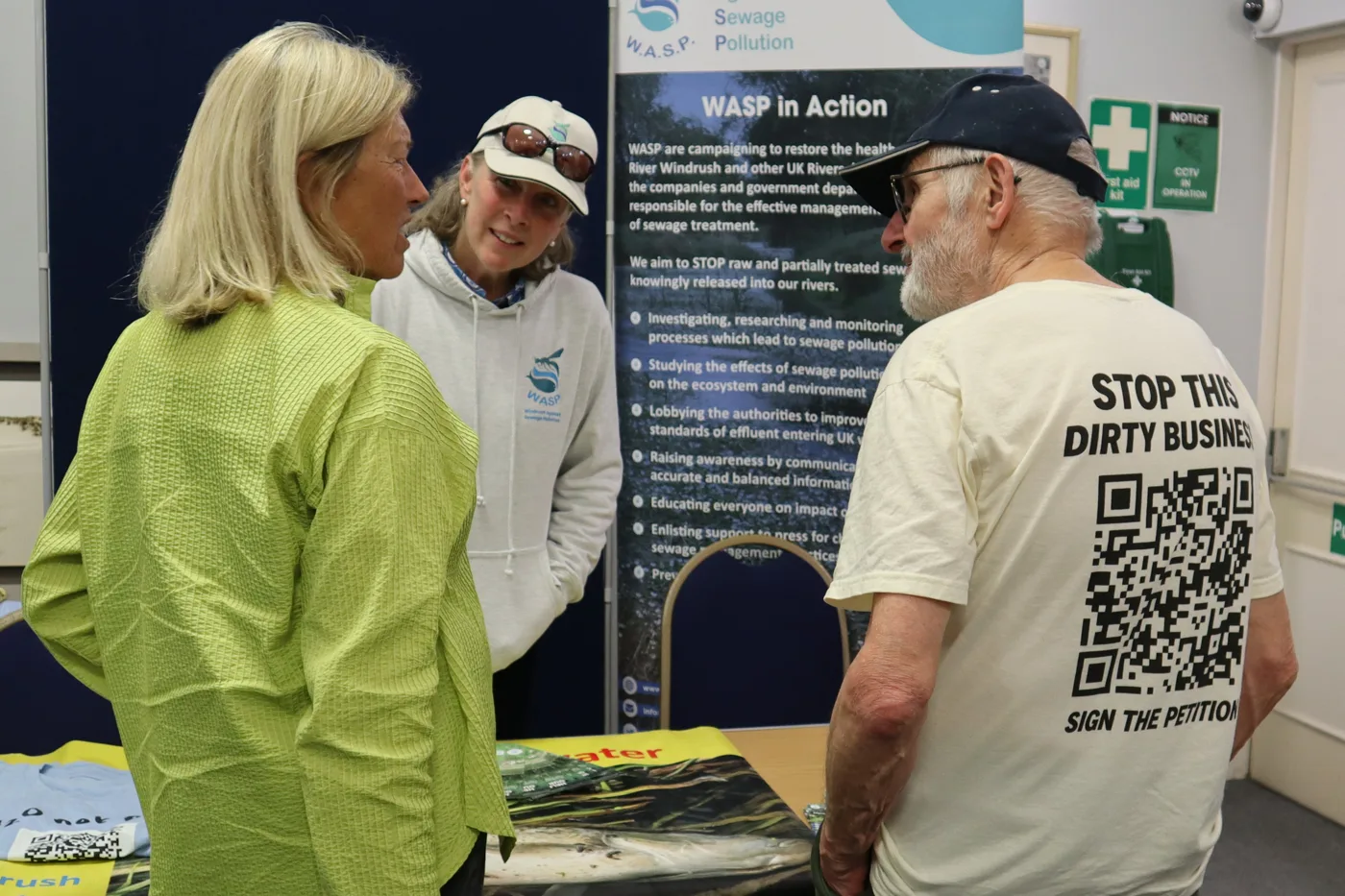 Visitors talking at the Windrush Against Sewage Pollution stand, in front of a WASP in Action banner; one man wears a T-shirt reading Stop this dirty business, sign the petition