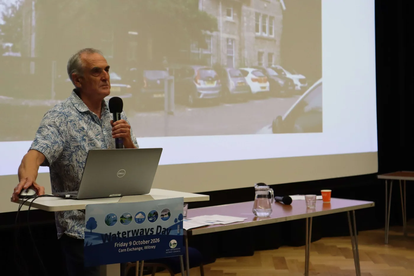 Ash Smith of Windrush Against Sewage Pollution speaking at a lectern marked Waterways Day, Friday 9 October 2026, Corn Exchange, Witney, in front of a projected photograph of a street