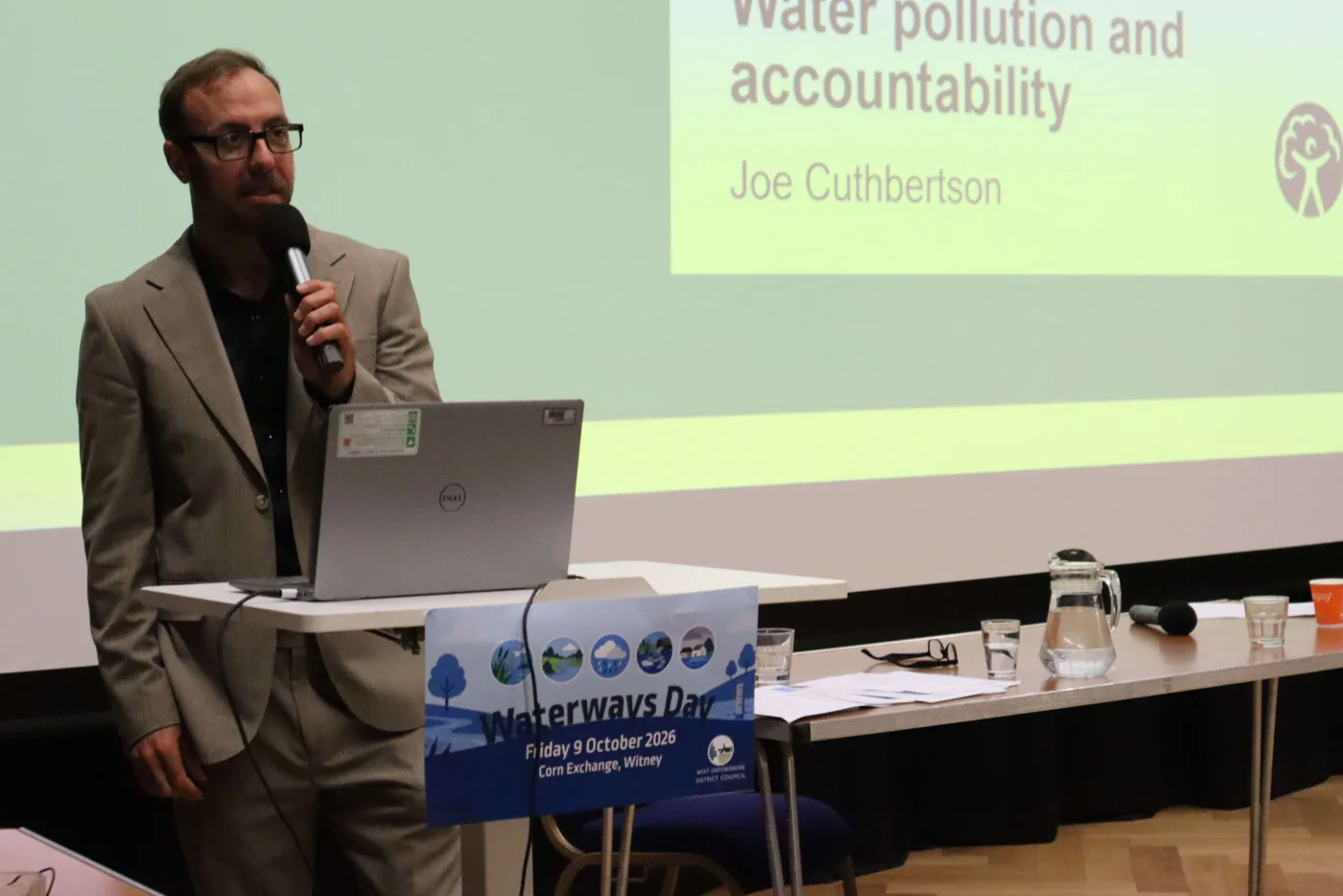 Joe Cuthbertson of the Environment Agency speaking into a microphone at the lectern, with a slide reading Water pollution and accountability behind him