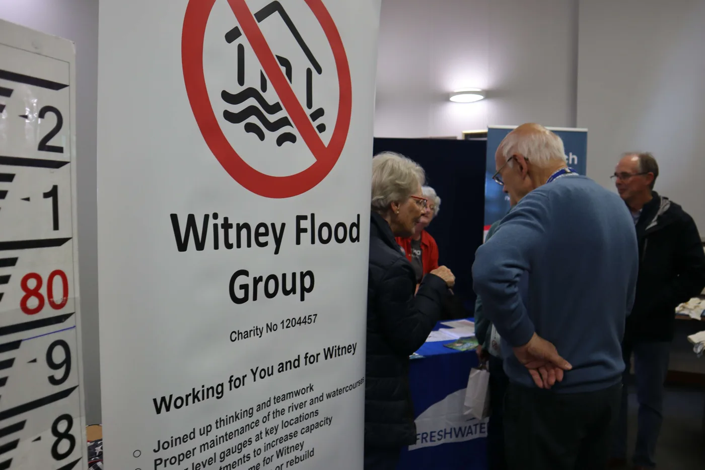 Visitors talking beside a Witney Flood Group banner and a large flood depth gauge marker