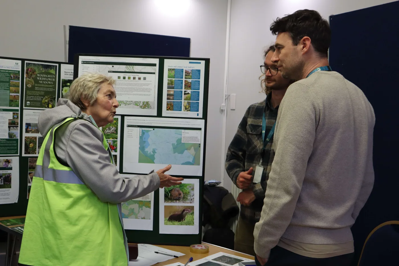 A woman in a high-visibility vest talks to two visitors in front of display boards of maps and wildlife photographs